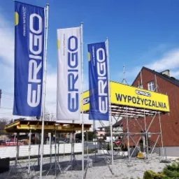 Flagi i żółty baner z napisem 'Wypożyczalnia' przed budynkiem z czerwonej cegły, widoczne rusztowania i elementy konstrukcyjne.