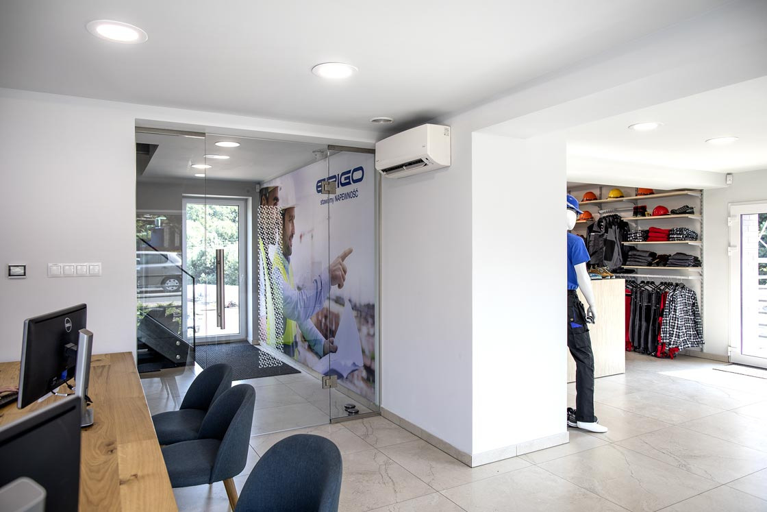 Wnętrze biura firmy budowlanej z recepcją, szklanymi drzwiami z grafiką budowlańców w kaskach i kamizelkach oraz ekspozycją odzieży roboczej na półkach i manekinie.