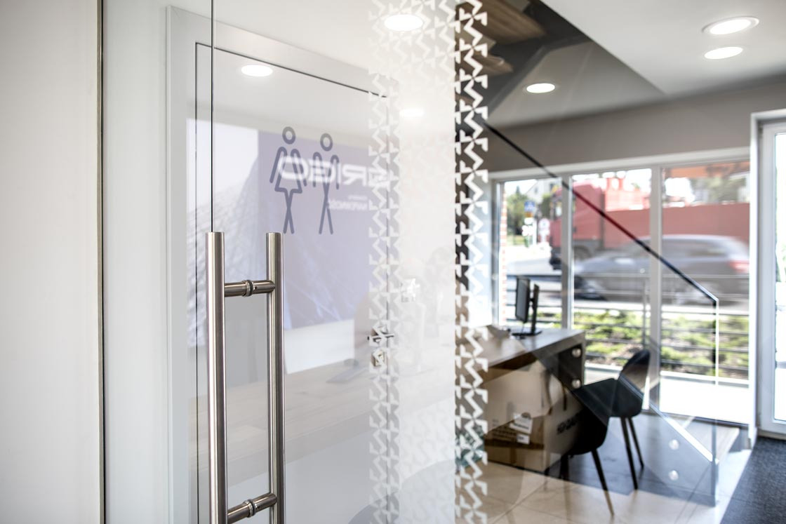 Szklane drzwi wejściowe z dekoracyjnym wzorem i uchwytem ze stali nierdzewnej w biurze w Radlinie, widok z bliska na wnętrze z biurkiem i krzesłem.