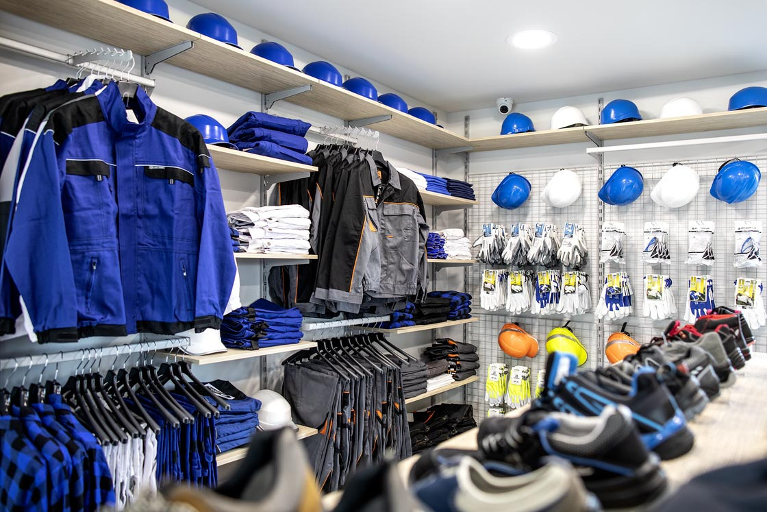 Sklep z odzieżą roboczą: niebieskie i szare kombinezony wiszą na wieszakach obok stosów spodni i koszul, na półkach kaski ochronne w różnych kolorach, a na ścianie rękawice robocze i buty ochronne.