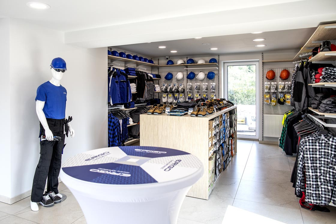 Wnętrze sklepu z odzieżą i akcesoriami roboczymi: Manekin w niebieskiej koszulce i czarnych spodniach roboczych z pasem na narzędzia, biały stół z logo, półki z butami roboczymi, kaskami i odzieżą...