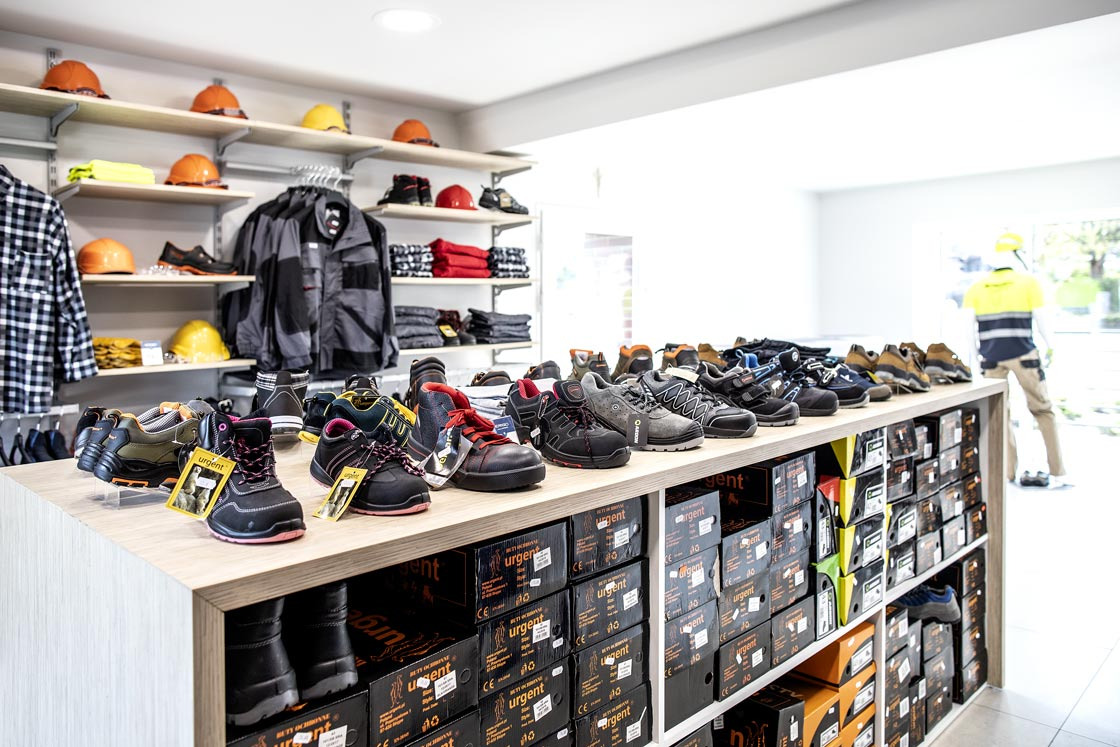 Ekspozycja w sklepie z obuwiem roboczym: różnorodne modele butów na półce, w tle kaski budowlane i odzież ochronna, a w oddali sylwetka pracownika w uniformie.