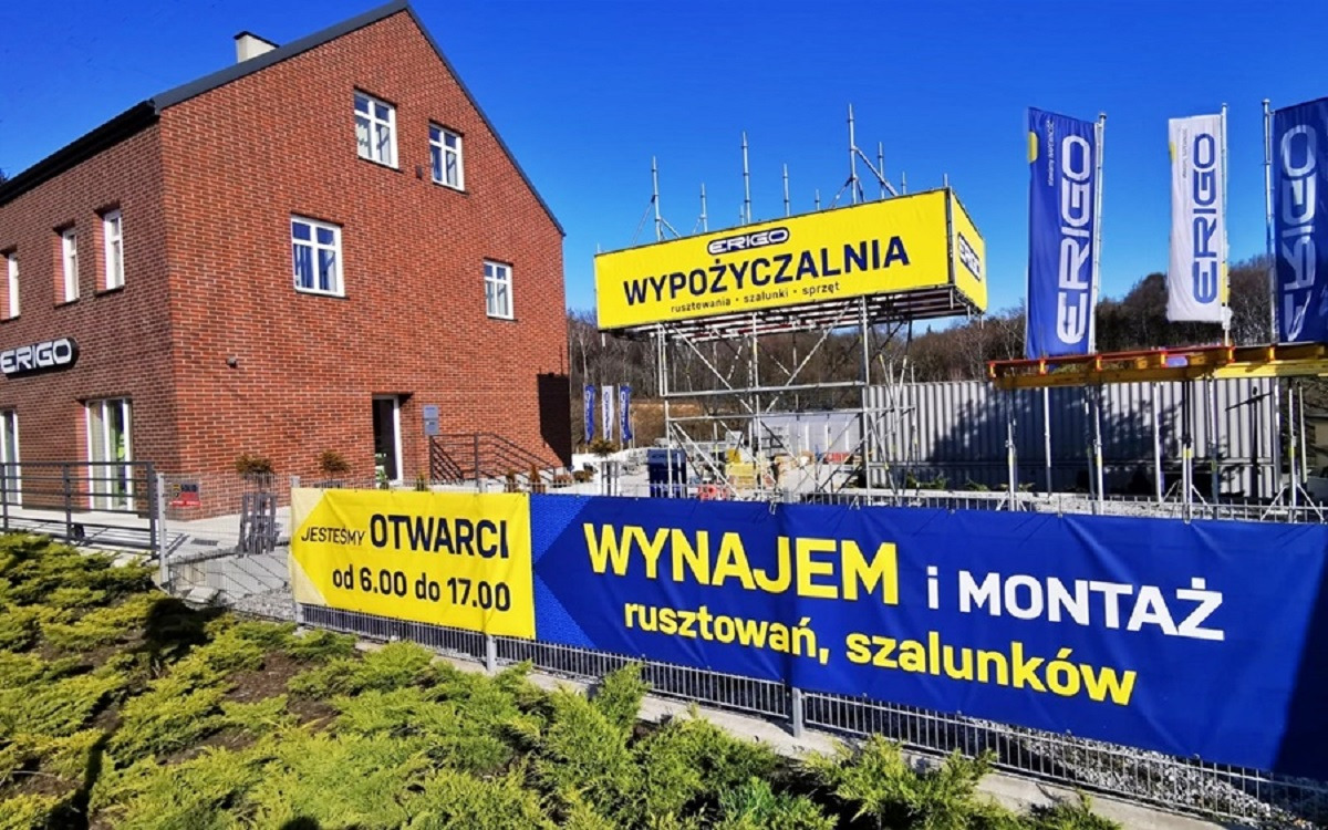 Budynek wypożyczalni sprzętu budowlanego z banerami reklamowymi oferującymi wynajem i montaż rusztowań oraz szalunków, widoczne logo firmy i godziny otwarcia.