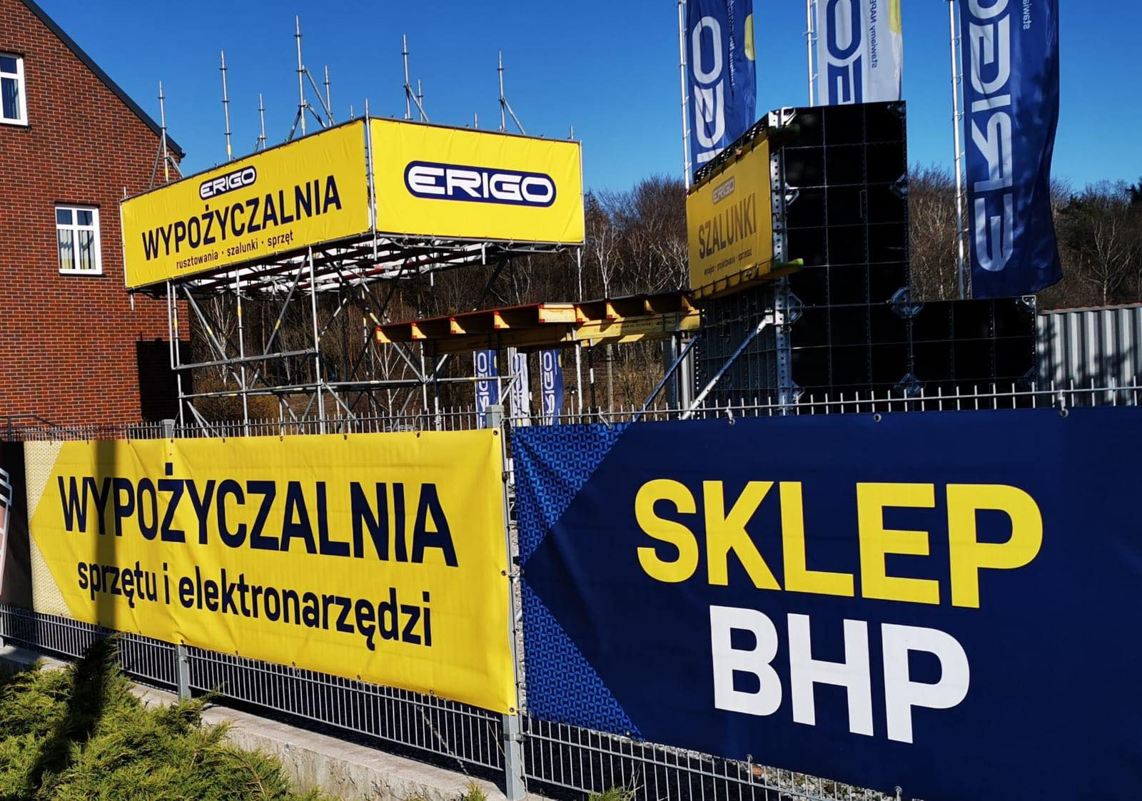 Żółte banery reklamowe z napisem 'WYPOŻYCZALNIA sprzętu i elektronarzędzi' oraz 'ERIGO WYPOŻYCZALNIA rusztowania szalunki sprzęt' umieszczone na ogrodzeniu i rusztowaniach przed budynkiem, obok...