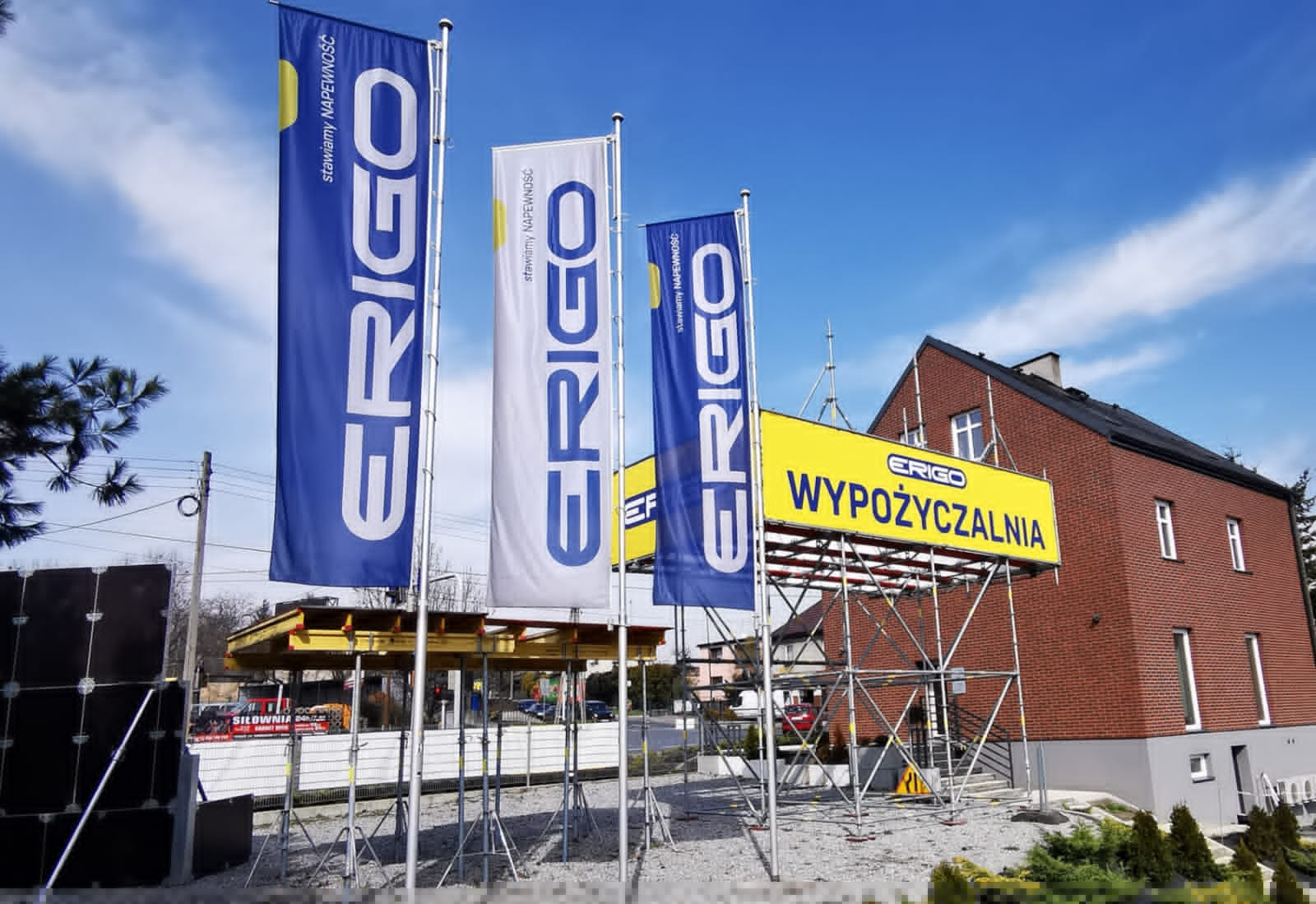 Flagi i żółty baner z napisem 'Wypożyczalnia' przed budynkiem z czerwonej cegły, widoczne rusztowania i elementy konstrukcyjne.