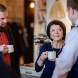 Konsultacja biznesowa przy kawie: kobieta w granatowej sukience uśmiecha się, trzymając biały kubek, podczas gdy dwóch mężczyzn w garniturach dyskutuje.