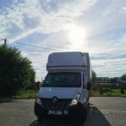 Biały bus dostawczy Renault Master z wysoką kabiną, zaparkowany na brukowanym placu, z włączonymi światłami mijania, na tle błękitnego nieba z chmurami.