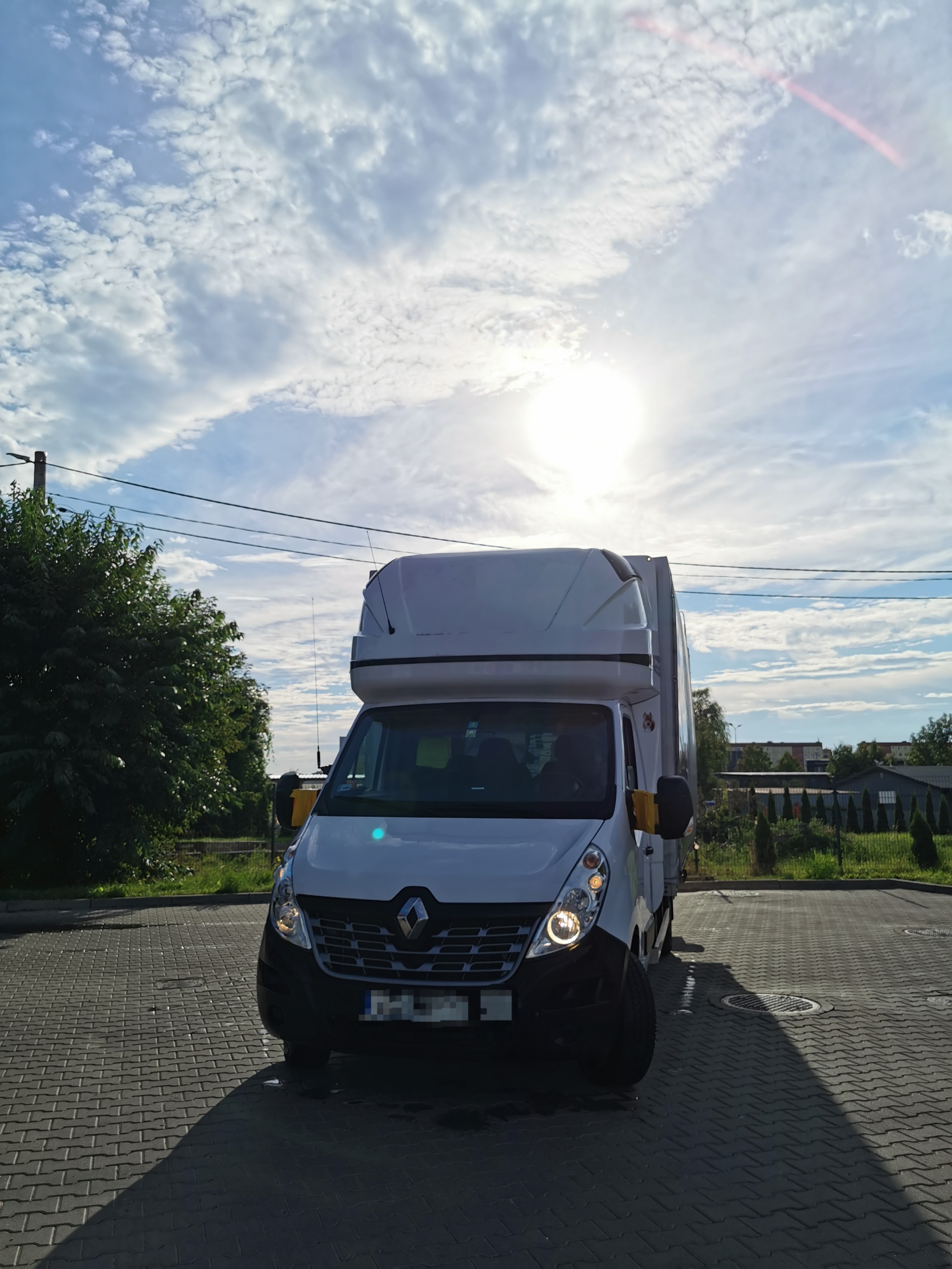 Biały bus dostawczy Renault Master z wysoką kabiną, zaparkowany na brukowanym placu, z włączonymi światłami mijania, na tle błękitnego nieba z chmurami.