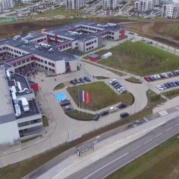 Przedsiębiorstwo Budowlane TB.INVEST Tomasz Brzeziński - Widok z lotu ptaka na nowoczesny, dwukondygnacyjny budynek szkoły z czerwonymi akcentami, otoczony parkingiem, zielenią i drogą dojazdową.