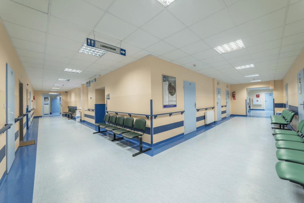 Jasny korytarz w placówce medycznej z zielonymi krzesłami dla pacjentów, tabliczką wskazującą laboratorium i niebieskimi akcentami kolorystycznymi na ścianach i podłodze.