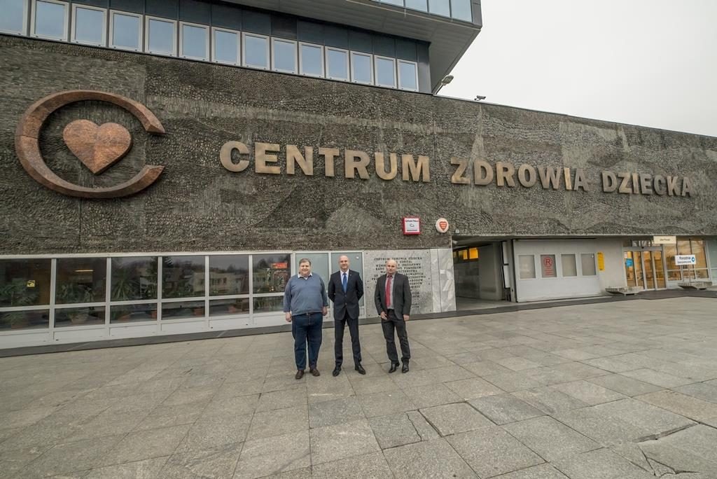 Trzech mężczyzn w garniturach stoi przed budynkiem Centrum Zdrowia Dziecka z charakterystycznym logo serca w okręgu. Fasada budynku wykonana z szarego, teksturowanego betonu. Widoczne okna...