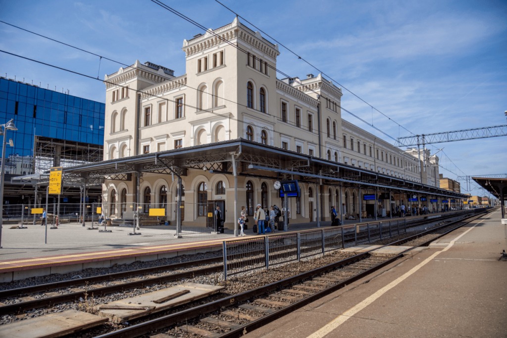 Odnowiony budynek dworca kolejowego z kremową fasadą i dwoma wieżami, widoczny z peronu z torami i oczekującymi pasażerami.