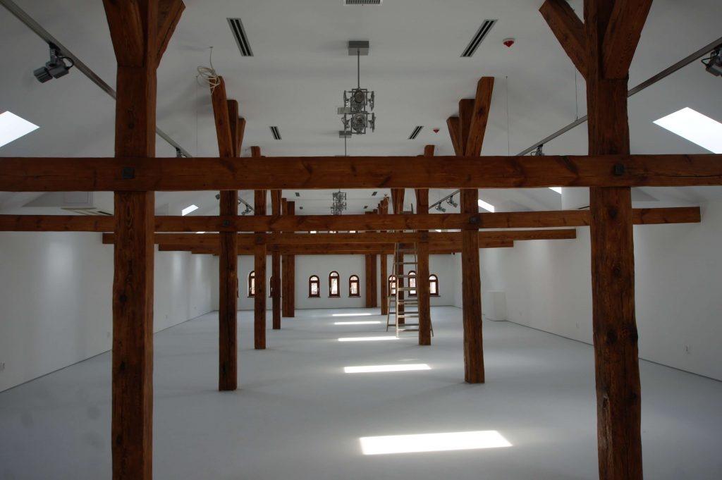 Przestronne wnętrze z drewnianą konstrukcją szkieletową, białymi ścianami i podłogą, oświetlone światłem wpadającym przez okna dachowe i rzędem małych okien na końcu sali, z widoczną drabiną opartą...