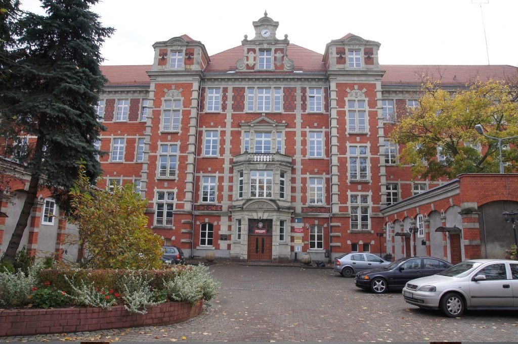 Elewacja odnowionego, trzypiętrowego budynku z czerwonej cegły z białymi obramowaniami okien, balkonem nad wejściem i zegarem na szczycie, otoczona zielenią i zaparkowanymi samochodami.