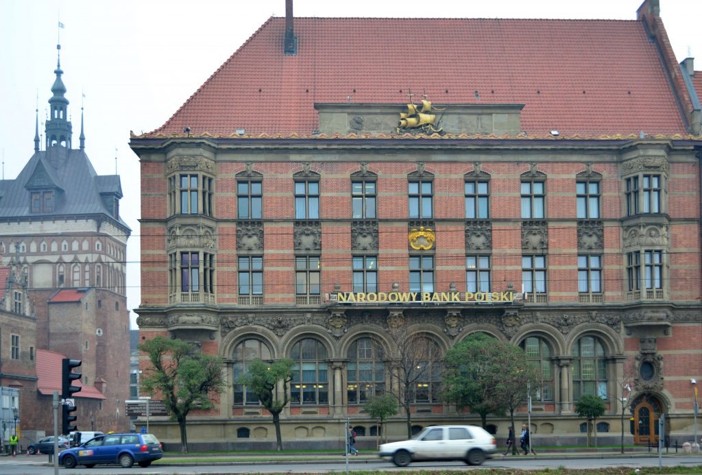 Fasada budynku Narodowego Banku Polskiego z czerwonej cegły, zdobiona ornamentami i rzeźbami, z widocznym napisem 'NARODOWY BANK POLSKI' oraz złotą statuetką żaglowca na szczycie, na tle szarego...
