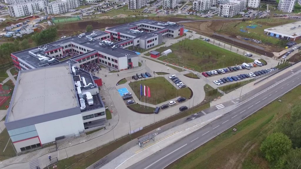 Widok z lotu ptaka na nowoczesny, dwukondygnacyjny budynek szkoły z czerwonymi akcentami, otoczony parkingiem, zielenią i drogą dojazdową.