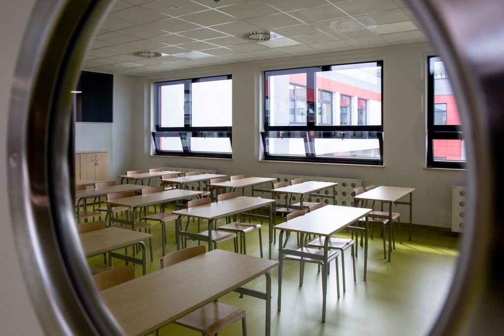 Pusta klasa szkolna z rzędami drewnianych ławek i krzeseł, widoczna przez okrągłe okno w drzwiach, z oknami wychodzącymi na budynek z czerwonymi panelami.