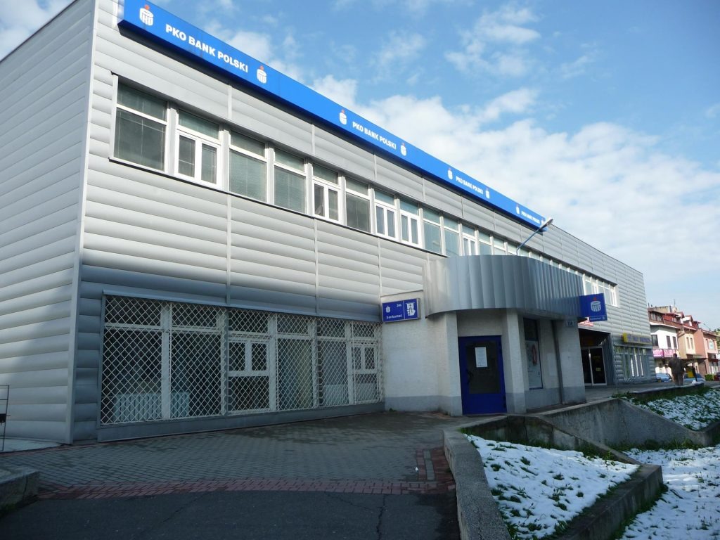 Budynek banku PKO BP z szarą elewacją z blachy falistej i niebieskim szyldem, widoczne okna z kratami i fragmenty topniejącego śniegu.