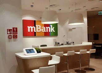 Wnętrze placówki mBanku: biała lada recepcyjna z czterema białymi hokerami, podwieszane białe lampy, logo mBank na ścianie, tablet na blacie z lewej strony, system przeciwpożarowy na ścianie po...