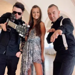 Trzyosobowy zespół muzyczny pozuje do zdjęcia: gitarzysta w czarnych okularach i z tatuażami, wokalistka w wzorzystej sukience i mężczyzna w czarnej koszuli z szelkami.