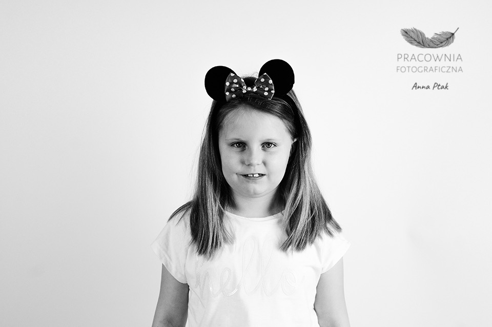 Czarno-biały portret dziewczynki z opaską Myszki Miki na głowie, na tle białej ściany, z logo fotografa w prawym górnym rogu.