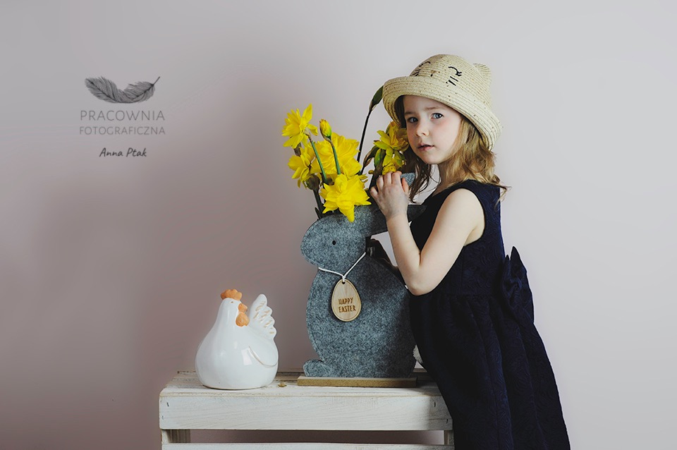 Dziewczynka w słomkowym kapeluszu z uszami kota, opiera się o szarego filcowego zająca z żółtymi narcyzami, obok ceramiczna biała kura na białej skrzynce.