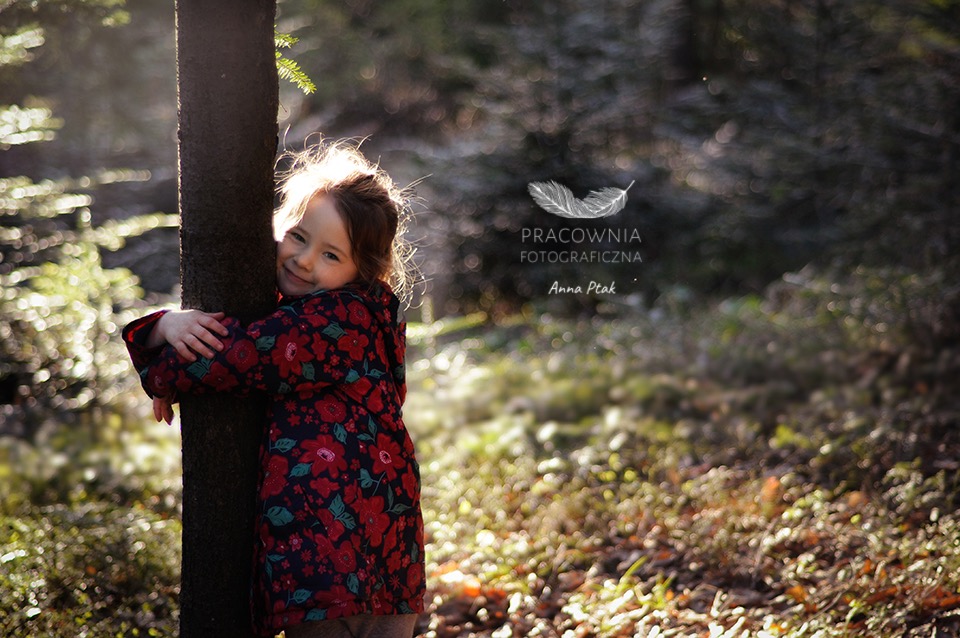 Dziewczynka w kwiecistej kurtce przytula się do drzewa w lesie, w tle rozproszone światło i logo fotografa.