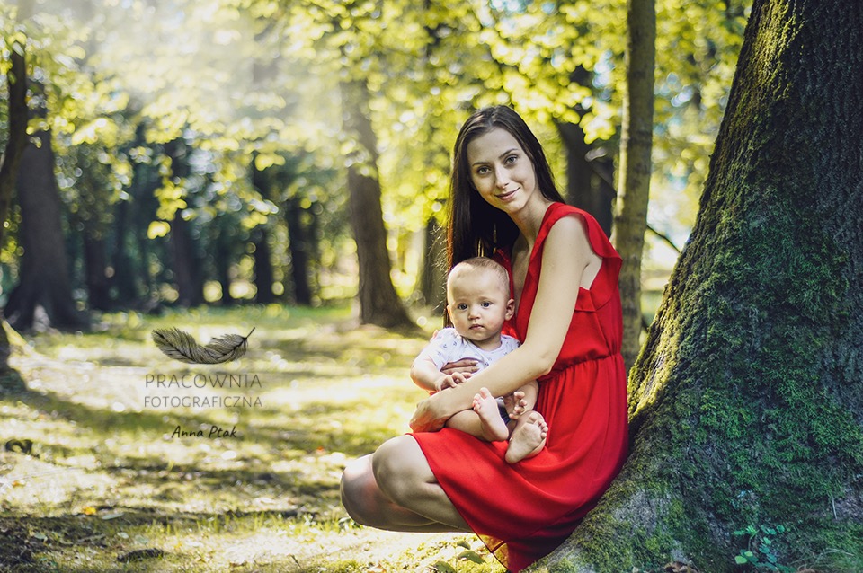 Mama w czerwonej sukience trzyma niemowlę na tle drzewa w lesie, w tle rozproszone światło słoneczne i logo fotografa w lewym dolnym rogu.