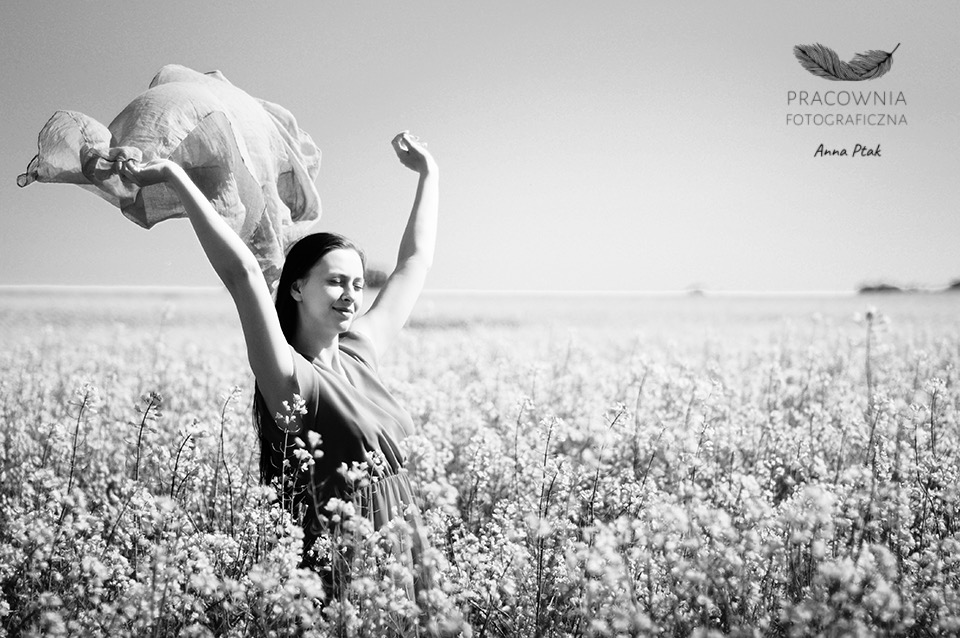 Czarno-białe zdjęcie kobiety w sukience stojącej na polu kwitnących kwiatów, trzymającej w górze zwiewny materiał, z logo fotografa w prawym górnym rogu.