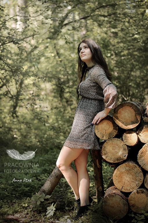 Młoda kobieta w sukience w panterkę opiera się o stos drewna na tle leśnym, spoglądając w górę podczas sesji fotograficznej.