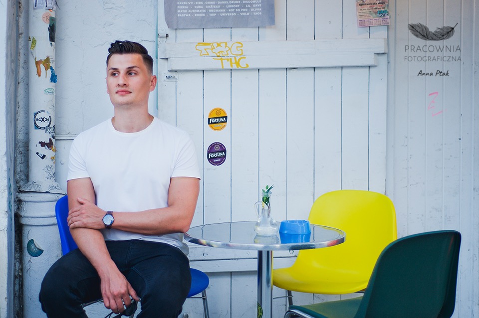 Mężczyzna w białym t-shircie siedzi na niebieskim krześle na tle białej ściany z naklejkami i graffiti, obok stolika z dzbankiem i kolorowymi krzesłami.