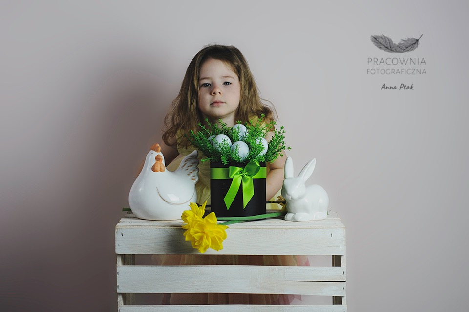 Dziewczynka z długimi włosami pozuje za kompozycją wielkanocną z ceramiczną kurą, zającem i pudełkiem z zielonymi gałązkami i białymi jajkami na białej skrzynce.