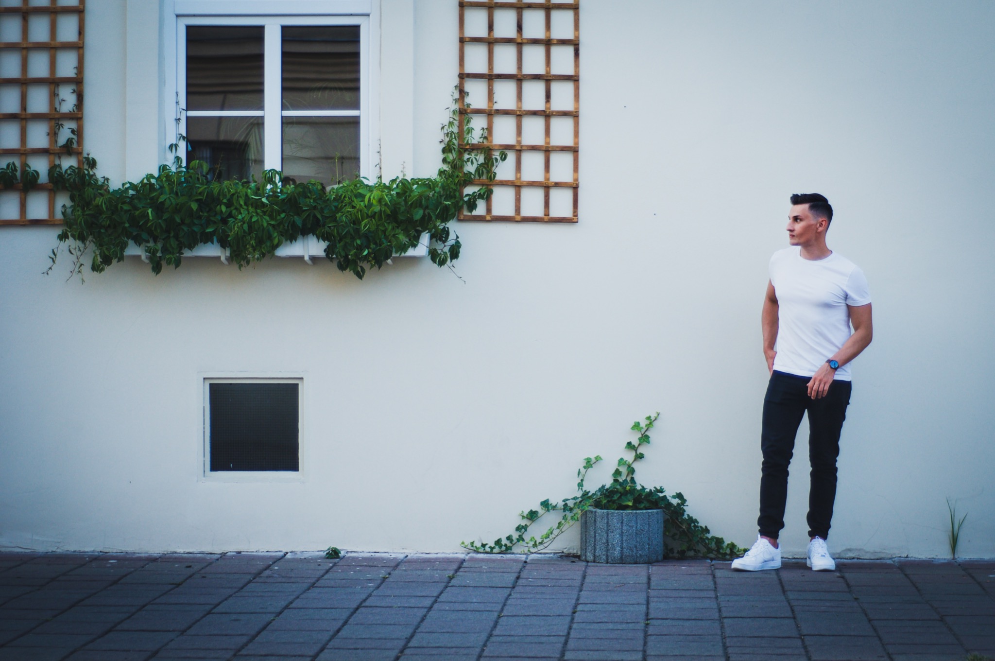 Mężczyzna w białym T-shircie i ciemnych spodniach stoi na tle jasnej ściany z oknami ozdobionymi zielenią i drewnianą kratką.