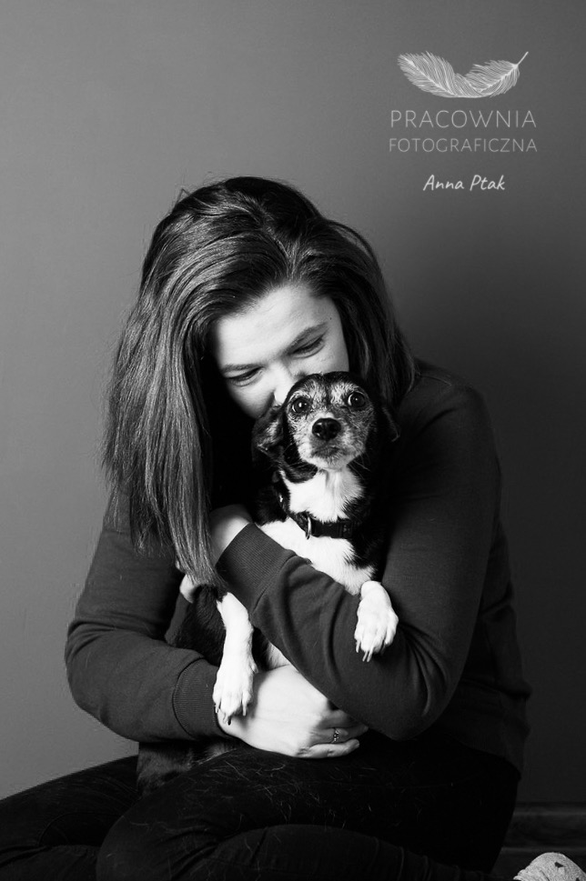 Czarno-białe zdjęcie kobiety obejmującej małego psa z obrożą; w tle logo pracowni fotograficznej z piórkiem.