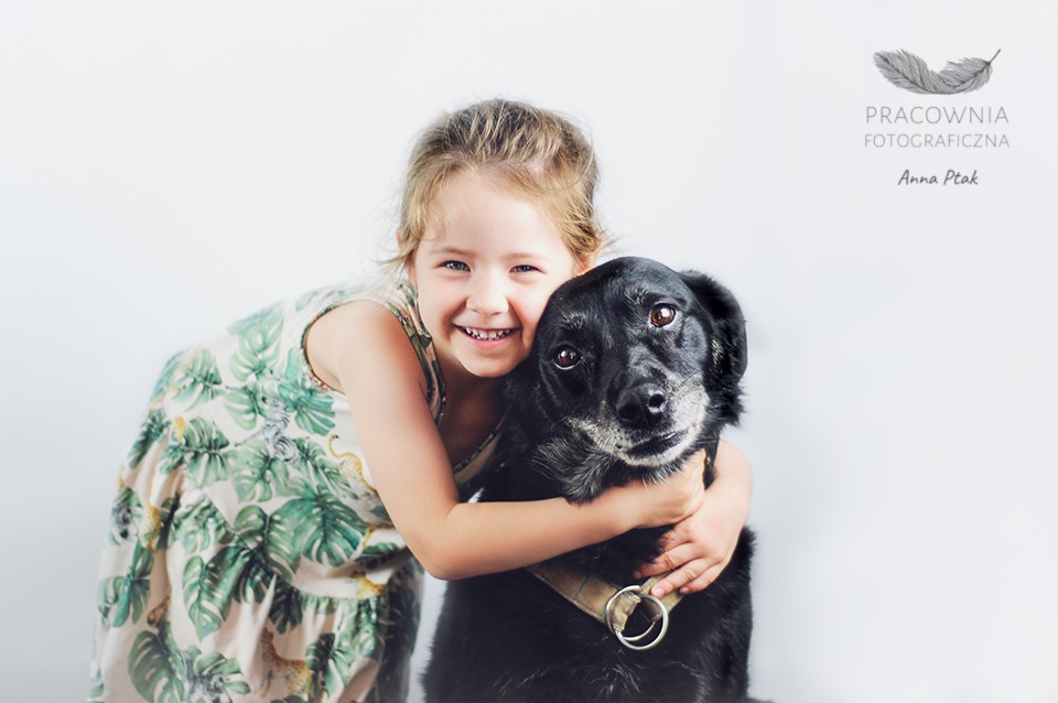 Uśmiechnięta dziewczynka w sukience z motywem tropikalnych liści przytula czarnego psa z szelkami na białym tle, w prawym górnym rogu logo pracowni fotograficznej.