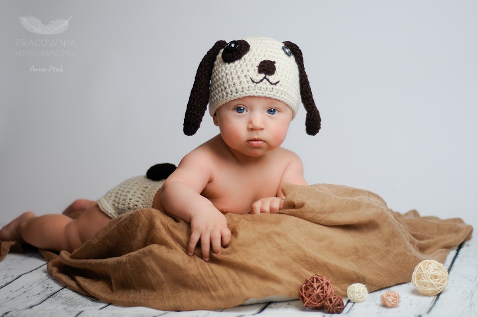 Niemowlę z niebieskimi oczami leżące na brązowym materiale, ubrane w dzierganą czapkę z motywem psa i majtki z pomponem, na białym drewnianym tle, z dekoracyjnymi kulami.
