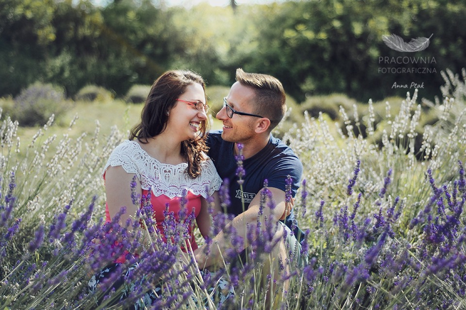 Uśmiechnięta para w okularach, siedząca w polu lawendy, z logo fotografa w prawym górnym rogu.