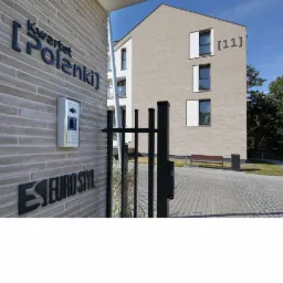 Nowoczesny budynek apartamentowy z elewacją z jasnej cegły, widoczny szyld z nazwą 'Kwartet Polanki' i numerem budynku '[11]', brama wejściowa i chodnik z kostki brukowej.