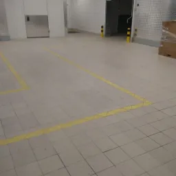 Pusta hala przemysłowa z jasnymi płytkami na podłodze, żółtymi liniami wyznaczającymi strefy, metalowymi panelami na ścianach i otwartą bramą wjazdową.