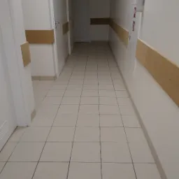 Pusty korytarz w budynku użyteczności publicznej z białymi ścianami, jasnymi płytkami na podłodze i brązowymi listwami ochronnymi na ścianach.