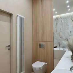 Pracownia Projektowa Dorota Wróbel - Nowoczesna łazienka z beżowymi ścianami, grzejnikiem żeberkowym, podwieszaną toaletą i drewnianą szafką. W lustrze odbija się wanna i marmurowe płytki.
