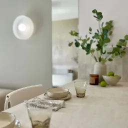 Minimalistyczna aranżacja stołu w jasnej tonacji: ceramiczne naczynia, szklanki, świeca i wazon z eukaliptusem na tle ściany z oświetleniem punktowym.