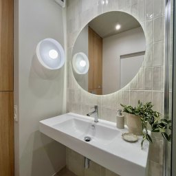 Pracownia Projektowa Dorota Wróbel - Nowoczesna łazienka z umywalką nablatową, okrągłym lustrem z podświetleniem LED i płytkami w odcieniach beżu. Roślina doniczkowa dodaje świeżości.