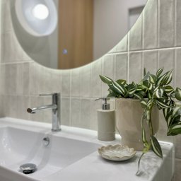Pracownia Projektowa Dorota Wróbel - Nowoczesna łazienka: biała umywalka z chromowaną baterią, okrągłe lustro z podświetleniem, ceramiczna doniczka z zieloną rośliną i mydelniczka w kształcie kwiatka.