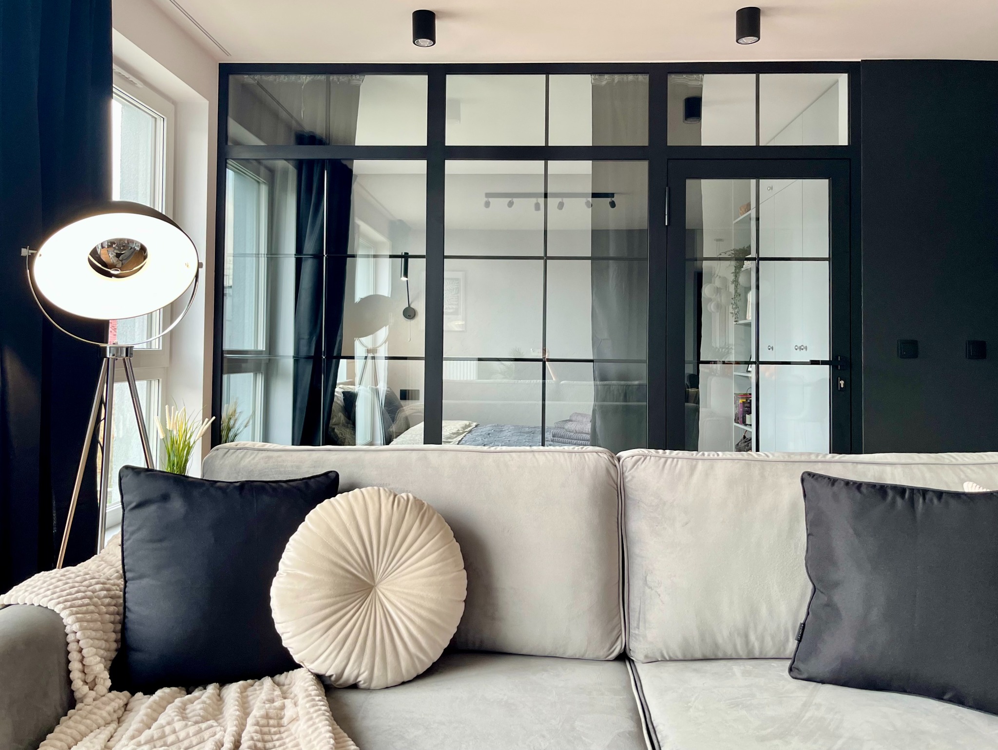 Salon w Krakowie z szarą sofą z poduszkami w odcieniach beżu i czerni, lampą podłogową na trójnogu i przeszkloną ścianą działową z czarnymi szprosami, za którą widać sypialnię.