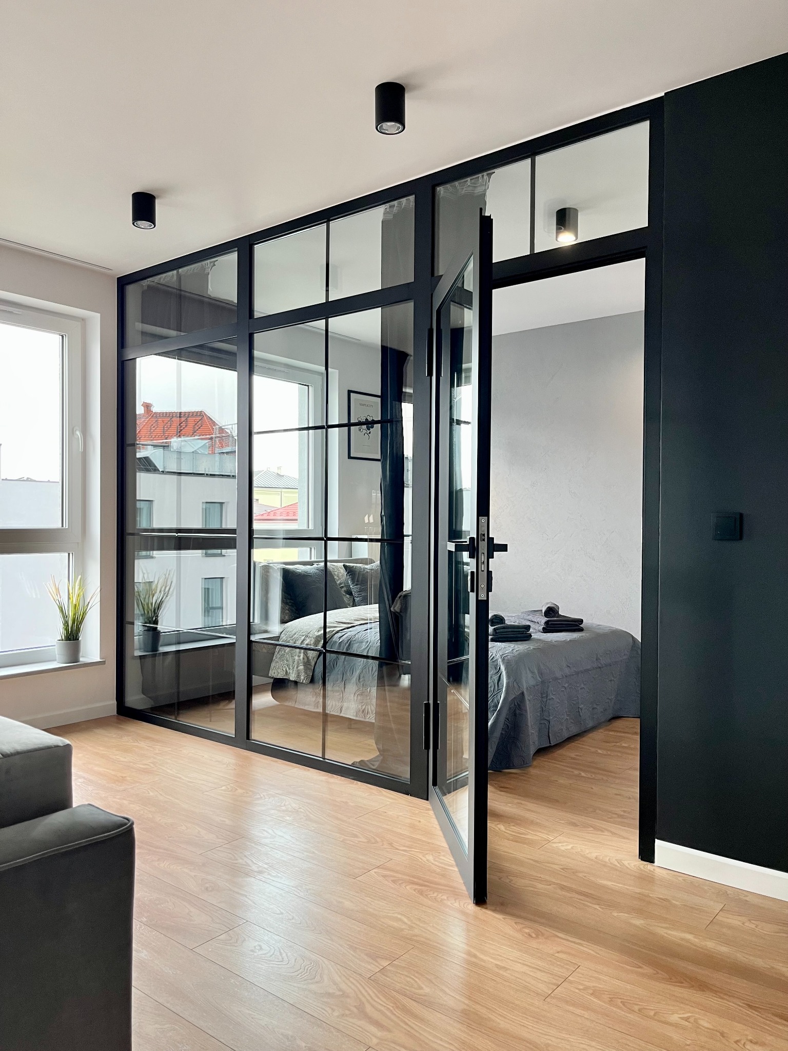Sypialnia oddzielona od salonu czarną, przeszkloną ścianą z otwieranymi drzwiami, widok na łóżko z szarą pościelą i ułożonymi ręcznikami.