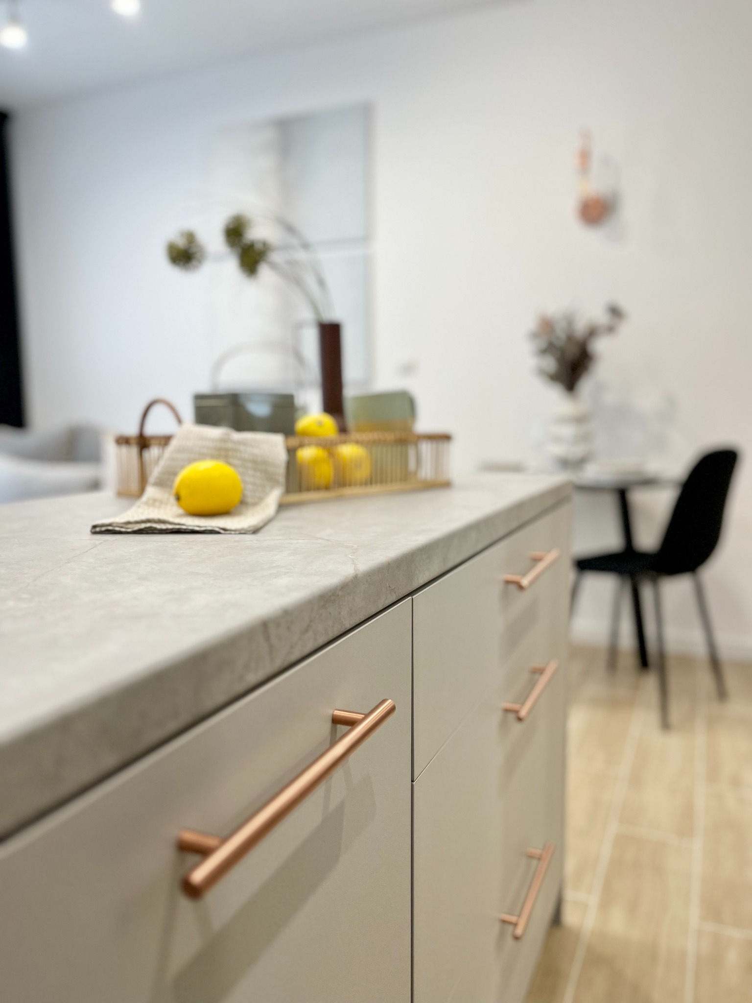 Blat kuchenny z szarego kamienia z miedzianymi uchwytami szafek, na blacie leżą cytryny na lnianej ściereczce, w tle stół z czarnym krzesłem i dekoracje.