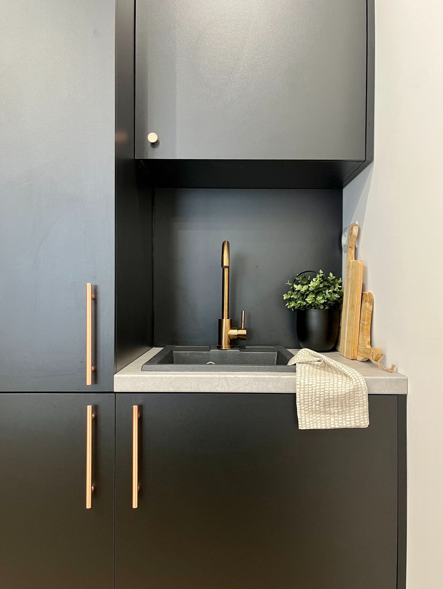 Nowoczesna, minimalistyczna kuchnia: czarne szafki z miedzianymi uchwytami, szary blat, złota bateria, czarny zlew i doniczka z zieloną rośliną, deski do krojenia i beżowa ścierka.