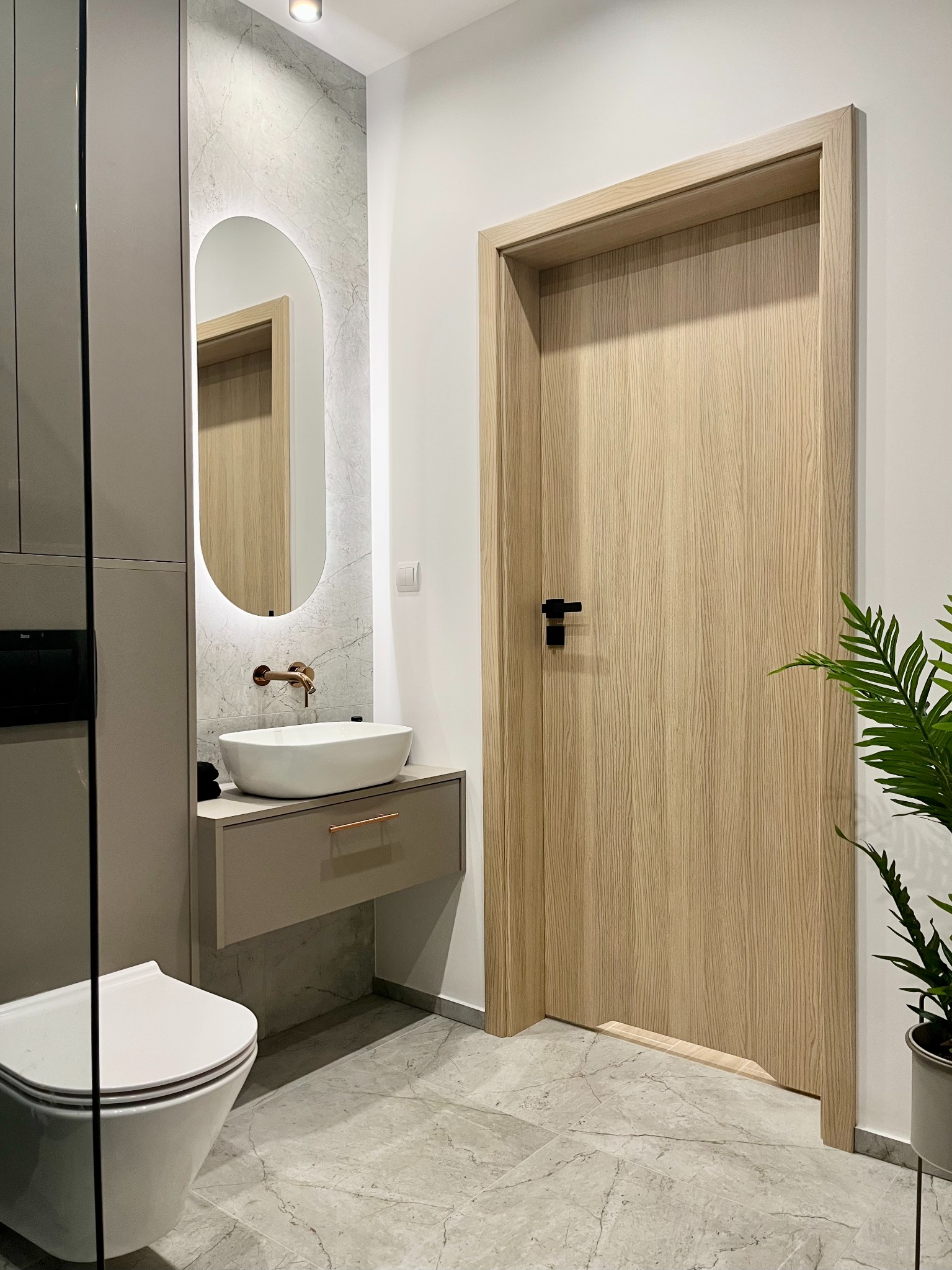 Nowoczesna łazienka z owalnym lustrem podświetlanym LED, umywalką nablatową na szafce w kolorze taupe, drewnianymi drzwiami z czarną klamką i marmurową podłogą.