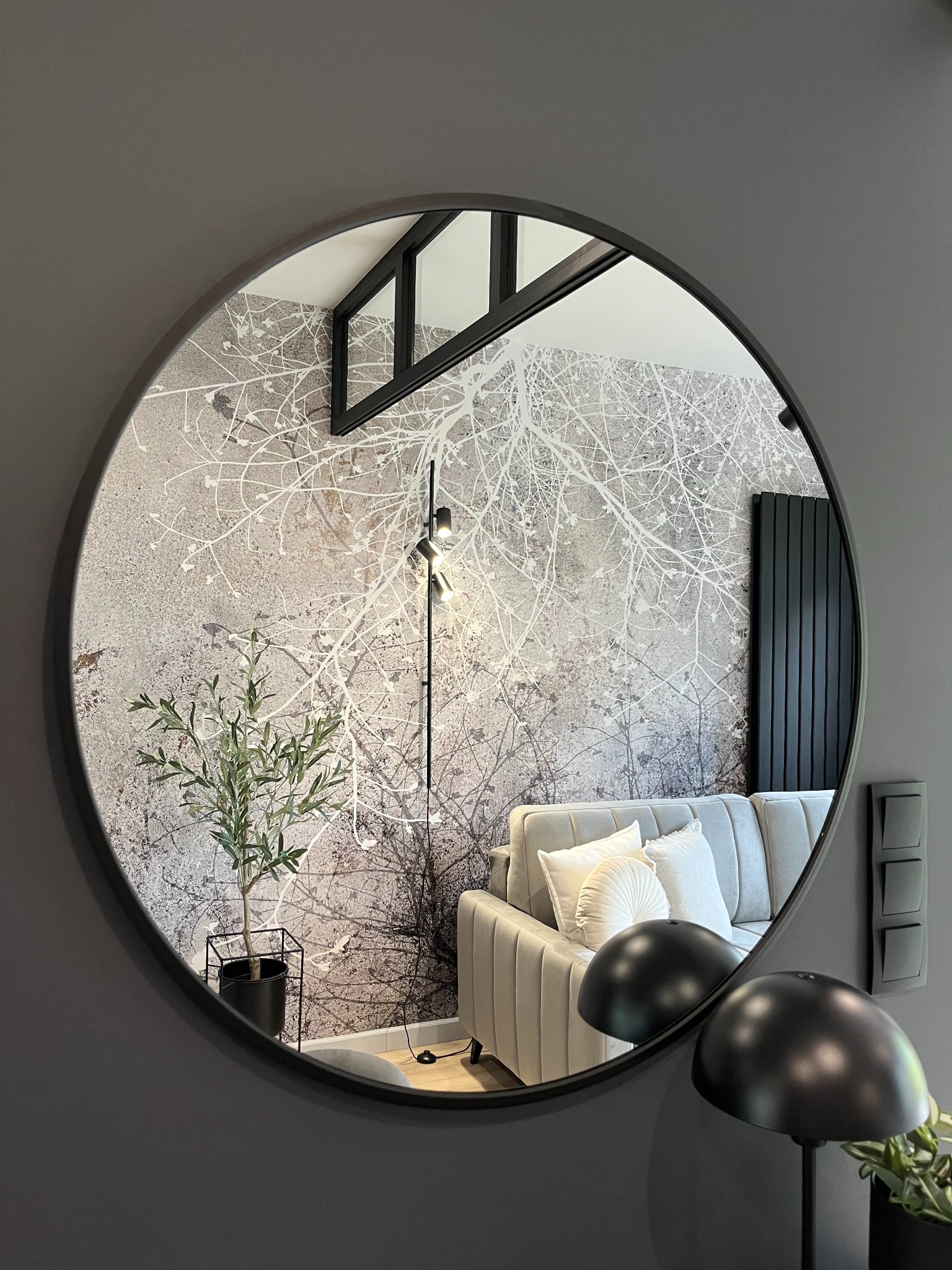 Okrągłe lustro odbija fragment salonu w odcieniach szarości z sofą, lampą podłogową i wzorzystą tapetą imitującą drzewo, uzupełnionego rośliną w czarnej doniczce.